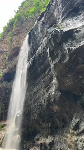 A powerful waterfall cascades down a sheer, dark rock face, surrounded by lush green vegetation clinging to the cliff above. The water is a bright white stream against the shadowy rock, creating a mist at the bottom. The rock face appears wet and textured, with visible strata and a rough surface. The overall impression is one of natural grandeur and the raw power of water in a rugged environment.