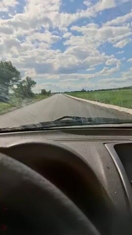 A first-person perspective video captures the view from inside a car driving down a straight, paved road surrounded by flat, green fields under a bright, cloudy sky. The steering wheel and dashboard are visible in the foreground. The camera is mounted inside the car, looking forward through the windshield. The road stretches out into the distance, with a clear horizon. The sky is a vibrant blue with scattered, puffy white clouds. The sun is shining, casting light on the landscape. There are no other vehicles or people visible on the road or in the surrounding fields. The scene appears to be a rural or agricultural area.