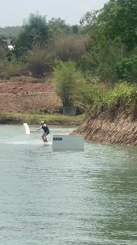 The video shows a man wakeboarding on a lake. He is wearing a life vest and helmet. He successfully rides over a floating rectangular platform. The lake is surrounded by green trees and a hillside. The water is a light blue-green color. Another floating platform is visible in the distance.