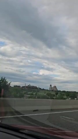 The video shows a view from a vehicle moving along a highway. In the foreground, the edge of a car's dashboard and windshield is visible, with a reflection of the interior on the glass. A concrete barrier separates the highway lanes. In the midground, there is a line of trees and some buildings on a hill. The most prominent structures on the hill are two large buildings, one with a domed roof and the other appearing to be a larger institutional building. A tall streetlight is also visible on the right side of the frame. The sky is cloudy with patches of blue sky peeking through. The overall impression is of a landscape viewed from a moving vehicle on an overcast day.