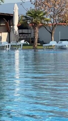 The video shows a poolside scene on a bright day. In the foreground, the surface of a swimming pool is visible, with clear blue water disturbed by gentle ripples. In the background, there are several white lounge chairs and umbrellas arranged around the pool. To the left, there's a large green tree with dense foliage. Further back, a building structure with a wooden element is visible, along with a tall palm tree to the right. The scene appears to be a resort or a private vacation spot. The camera is positioned low, likely at water level, looking towards the pool area and the surrounding landscape.