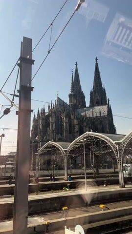 The video shows a view from a train as it passes through a station. In the background stands the Cologne Cathedral, a large Gothic-style church with two prominent spires. In the foreground is the train station platform, covered by a series of arched metal and glass canopies. There are a few people visible on the platform, some standing and others walking. A white service vehicle is parked on the platform. The sky is clear and blue.