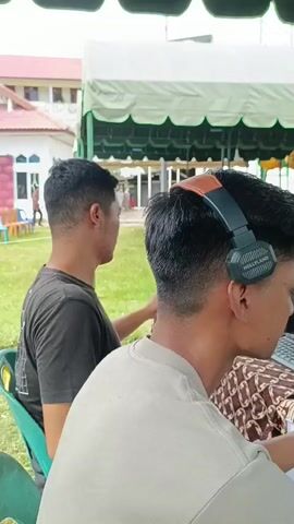 The video shows an outdoor event under a tent. In the foreground, two men are seen from the back. The man on the right wears a light-colored t-shirt and headphones, looking towards a computer monitor which displays various graphics. The man on the left wears a dark t-shirt and is facing away from the camera. Behind them, an audience is seated on chairs, facing a stage. A person is speaking at a podium on the stage, with a camera positioned in front of them. The environment appears to be a grassy area in front of a building.