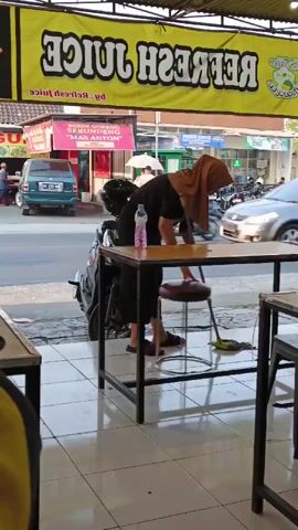 A woman wearing a brown headscarf and black clothing is mopping the floor with a green mop. She is standing next to a black motorcycle parked under a table. A bottle of water sits on the table. In the background, traffic moves along a street, including cars and motorcycles. A yellow banner with the words "REFRESH JUICE" is visible above the scene. The overall environment appears to be an outdoor seating area of a food establishment.