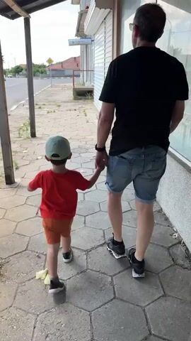 A father and his young son walk hand in hand down a sidewalk past a closed storefront. The father, wearing a black t-shirt and denim shorts, walks on the right, while the son, in a red t-shirt and orange shorts, walks on the left. The son wears a light blue baseball cap. They are walking away from the camera. The sidewalk is paved with hexagonal concrete tiles. A street with a yellow diamond-shaped sign is visible to the left of the sidewalk. The storefront to the right has closed metal shutters and a window. The sky is overcast.