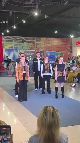 A group of models walks down a runway in a brightly lit indoor setting, likely a mall. The models are showcasing a collection of clothing that features orange and black patterns. In the foreground, a person's head and shoulders are visible as they hold up a smartphone to record the event. The background reveals a crowd of spectators seated and standing, watching the fashion show. Behind the runway, a large mural or backdrop depicts an urban landscape with construction elements. Storefronts, including one with a "zolla" logo, are also visible in the distance. The overall atmosphere appears to be a public event, possibly a fashion show or a cultural presentation.