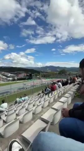 The video shows a person seated in stadium-style seating, seemingly at a racetrack. The sky is blue with scattered clouds. The person is wearing jeans and sneakers and holding their hands together in their lap. They appear to be looking at the track.

As the camera pans to the left, the view shifts to the racetrack. A bright yellow racecar speeds along the track. The track has a green and red painted curb on the inside edge.

The camera continues to pan, showing a wider view of the track as the racecar curves along the course. Other racecars can be seen further in the distance.

The camera also captures other spectators in the stands, some sitting and watching the race. In the background, the scenery consists of trees and buildings, giving the impression of a location in the countryside. There are signs on the fence.