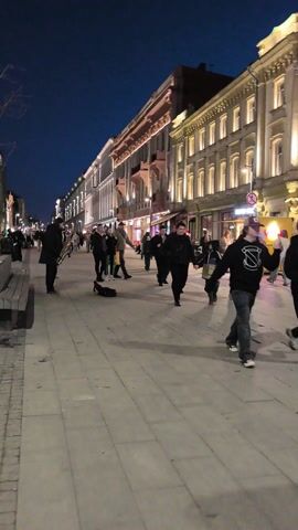 The video shows a street scene at night. The street is wide and paved with light-colored stone. People are walking in both directions. On either side of the street are tall buildings with illuminated windows. The sky is a deep blue. A man stands on the left side of the street playing a saxophone. A saxophone case lies on the ground near him. A few people are seated on benches nearby. The street is well-lit with streetlights and lights from the buildings.