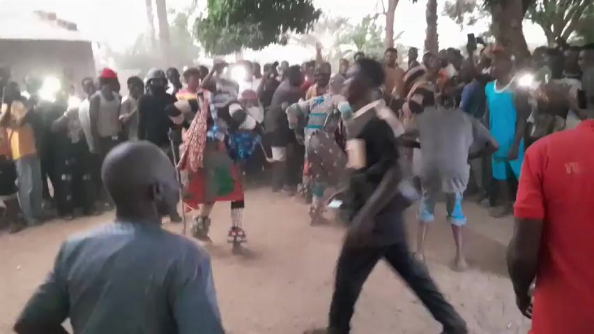 The video shows a large group of people gathered outdoors, possibly for a celebration or cultural event. Many are holding up cell phones, using the flashlight function to illuminate the scene. Several individuals, some dressed in colorful, traditional attire, are dancing and moving energetically. The background shows trees and simple buildings, suggesting a rural or village setting. The overall atmosphere appears lively and festive.