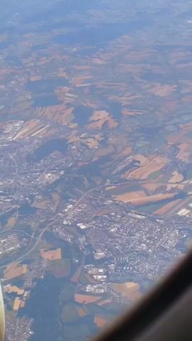 This video is an aerial view from an airplane window looking down at a landscape. The landscape is a mix of urban areas and agricultural fields. The urban areas appear to be towns or cities with buildings and roads. The agricultural fields are a patchwork of different colors, suggesting various crops or stages of growth. The sky is visible at the top of the frame, and a portion of the airplane's wing or fuselage is visible in the lower right corner. The scene is bathed in natural light, suggesting it is daytime. The camera is tilted, giving a sense of movement or perspective from the aircraft.