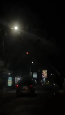 The video shows a night scene from inside a vehicle. The camera is pointed forward, capturing the road ahead and the dark sky. A faint orange moon is visible in the upper center of the frame, with a bright light source, possibly a streetlamp, above and to its left. The road is lined with buildings on either side, some of which have illuminated signs. A car is driving in front of the vehicle from which the video is recorded. The overall atmosphere is dimly lit and somewhat hazy.