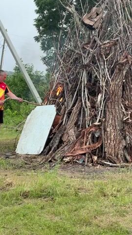 A man is shown using a stick to push a white board into a large bonfire. The bonfire is made of logs and branches, and there is smoke coming from it. The man is wearing a blue shirt and shorts. The background shows trees and a utility pole. The ground is covered in grass.