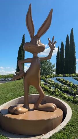 The video shows a golden Bugs Bunny statue in a park. Bugs Bunny is in a playful pose, with one hand raised making a peace sign. He is standing on a circular base, surrounded by green grass and a floral arrangement in the shape of the Warner Bros. logo. Tall cypress trees are visible in the background under a blue sky with scattered white clouds. The video pans from the statue to the floral logo.