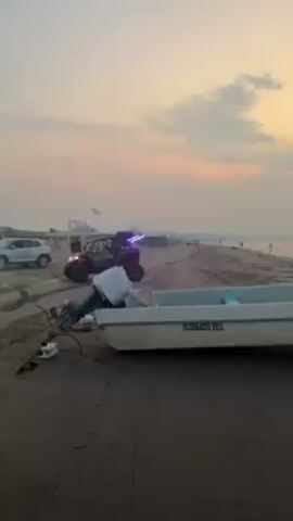 The video shows a sandy beach at dawn or dusk. The sky is a mix of light pink and gray clouds. A white boat with "ALVARADO, VER." written on its side rests on the sand in the foreground, along with some logs. Further down the beach, a red off-road vehicle with lights is parked. People are walking on the sand near the vehicle. A silver SUV is also parked in the background. A man in a red shirt passes by in the foreground, followed by a woman in a dress who stretches her arms out toward the ocean.
