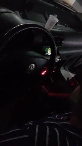Inside a parked car at night, a person with a beard and a striped shirt is seated in the passenger seat. The focus is on the Volkswagen steering wheel with its logo and illuminated red lights. A white plastic cup rests on the dashboard. The car's interior is dark, but the dashboard lights are visible. The door appears to be ajar. There's audio playing, featuring a male voice. The camera moves up to capture a partial view of the exterior of the parking garage.