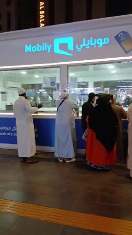 A group of people are standing outside of a Mobily mobile service shop, waiting for service. Most of the men are wearing traditional Muslim white robes and head coverings.  A woman is wearing an orange dress and a black head covering. The shop is brightly lit, and the Mobily logo is prominently displayed.  The building across the street has the words "DIYAR ALSALA" written vertically.