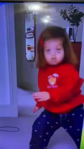 A young girl with brown hair and a red bow is dancing in a hallway. She is wearing a red sweatshirt with a bear design and navy blue patterned pants. Behind her is a white doorframe, a mirror, and a dark wooden cabinet with a plant on top. The lighting in the hallway appears to be from overhead lights. The video quality is somewhat low, and the colors are slightly washed out.