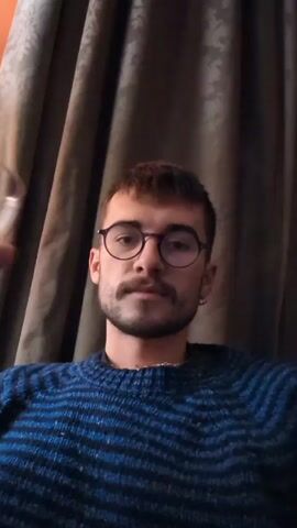 A person is drinking red wine from a wine glass. They are wearing glasses, a blue sweater, and a small earring. They have short brown hair and a mustache. A curtain is visible in the background. Voices are audible in the background.