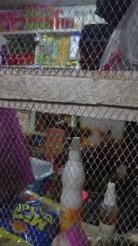 The video shows the interior of what appears to be a small shop or store. Shelves stocked with various goods, including food items and bottled drinks, line the walls.  A wire mesh partition partially obscures the view, suggesting a caged or sectioned area. The camera moves to reveal a man wearing traditional attire, including a patterned cap and a brown shirt. He is standing near the counter of the shop. The environment looks modest and suggests a casual, perhaps rural, setting.