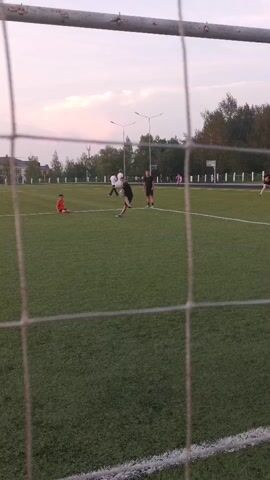 The video shows a group of children playing soccer on an artificial turf field. The camera is positioned behind a soccer net, looking out onto the field. In the foreground, a young boy with blond hair, wearing a white t-shirt and black shorts, stands in front of the goal. He appears to be acting as the goalkeeper. He is holding a soccer ball. A soccer ball is in motion, and it is apparent that the goalkeeper has just made or is about to make a save. In the background, several other children are visible on the field, some of whom are kicking the soccer ball. The sky is overcast with a pale pink hue, suggesting it is either early morning or late evening. There are trees and stadium lights visible in the distance, indicating the presence of a sports complex. The field is marked with white lines, typical of a soccer field.