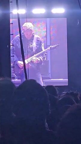 The video shows two different band members performing on stage, as seen on a large screen at a concert. The first clip features a guitarist wearing a dark jacket over a patterned shirt, playing a dark-colored electric guitar. The second clip shows a singer, who is wearing glasses and a dark shirt, singing into a microphone. They are in front of a drum set, and the stage lighting is purple. The angle of the video suggests it was recorded by someone in the audience. The lighting is bright on the stage.