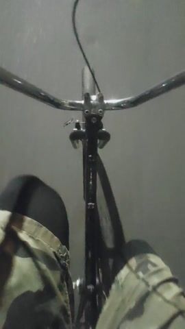 A first-person perspective video shows a person riding a bicycle. The camera is positioned low and facing forward, capturing the handlebars, the front wheel, and a portion of the rider's legs clad in camouflage pants. The background is a blurry, gray, concrete surface, possibly a street or sidewalk, with some indistinct objects and lights visible in the distance, suggesting an urban or semi-urban environment. The video appears to be shot at night or in low-light conditions, as indicated by the illumination from streetlights or vehicle headlights. The camera is slightly tilted, giving a sense of motion and movement as the bicycle is being ridden.