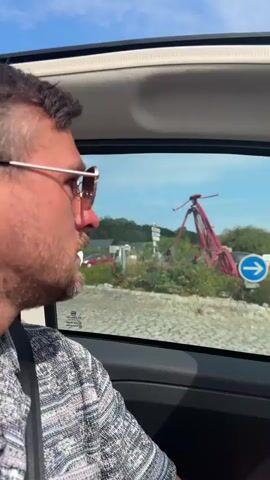 A man wearing sunglasses and a patterned shirt is in the passenger seat of a car, looking out the window. The car is moving and passing a roundabout. In the center of the roundabout is a large red sculpture of a bicycle. A blue directional sign with an arrow pointing to the right is also visible. The car then exits the roundabout and continues down a road. The driver's arm is visible on the steering wheel, and the Fiat logo is on the steering wheel. Buildings and trees line the road, and there are other cars on the road. A sign for "Weldon" is visible on a building on the right side of the road. The sky is blue with some clouds.