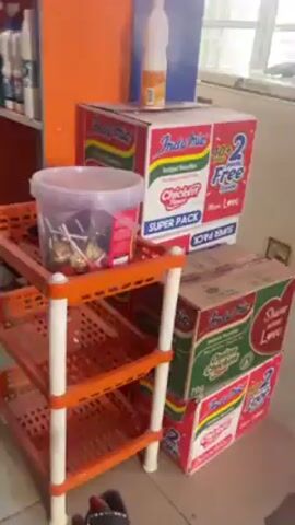 The video shows the interior of a small business space.  Several boxes of Indomie noodles are stacked near an orange shelving unit. Beside them, a stack of bags of rice is visible, topped with a package of diapers.  Miscellaneous objects, including what appears to be a tape measure and an orange cap, are scattered on the floor. The floor tiles are light colored with some staining. A white refrigerator is in the background.  Loud music with a distinctive electronic beat is playing.
