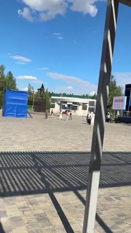 The video shows a paved public square on a sunny day. The square has a statue of a figure on horseback, a blue tent-like structure, and a stage on one side. Several people are walking across the square in various directions. The background includes a building with green accents on its roof. The foreground features the shadow and part of a metal structure. The sky is blue with scattered clouds.