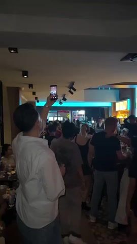 The video begins showing a crowded indoor venue, possibly a bar or club. In the foreground, a person from behind is holding up a smartphone to record the scene. The camera is angled upwards, capturing a lively atmosphere with many people gathered. Some are dancing or socializing. The venue has a ceiling with spotlights and a decorative element that glows with blue light. Further back, a bar area is visible with shelves stocked with bottles. Several individuals are interacting, and some appear to be staff members behind the bar. The lighting is dim, with colored lights contributing to the ambiance. The video continues to pan slightly, showing more of the crowd and the general activity within the venue.