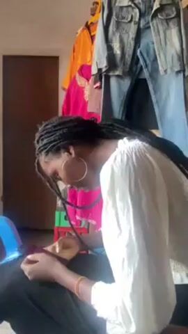 A woman with black dreadlocks and large hoop earrings is sitting on a blue stool, focused on working with fabric. She is wearing a white blouse.  Behind her, clothes hang on a rack, including a yellow top, a pink top, and a denim jacket. The room appears to be a workspace or small shop.