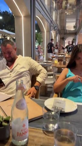 The video begins with a medium shot of a man and a woman seated at a table in what appears to be a restaurant. The man is wearing a light-colored shirt and looking down. The woman is wearing a turquoise top and looking at her phone. A bottle of San Pellegrino water and glasses are on the table. The background shows other diners and the restaurant's interior, with arched doorways and ambient lighting. The camera then pans up to a reflection on the ceiling, showing a busy restaurant environment with people dining and staff moving around. The video continues with a series of overhead shots of the same restaurant. The first overhead shot shows four people seated at a table, each looking at their phones or menus. The second overhead shot is similar, showing the same group of people at the table, with one person looking up. The subsequent overhead shots continue to show diners at tables, with some people actively engaged with their phones. The environment appears to be a modern restaurant with concrete floors and wooden tables. The lighting is dim, suggesting an evening setting.