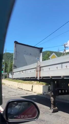 The video opens facing a road with a white truck parked on the right side, obscuring most of the view. The camera is inside a vehicle, looking out. A white concrete barrier runs along the side of the road. In the background, there are houses and a lush green hill under a bright blue sky. A pedestrian crossing is visible in the foreground. As the video progresses, the camera moves forward, revealing more of the road and surroundings. A stop sign is visible on the left. The truck then begins to move, turning across the intersection. The houses in the background are of various styles and colors, with fences and trees. Power lines crisscross the sky. The overall impression is a sunny day in a rural or semi-rural area.