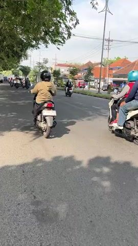 The video shows a view from a moving motorcycle on a paved road. In the foreground, two motorcyclists wearing helmets are riding away from the camera. The motorcyclist on the left is wearing a gray hoodie and blue jeans, while the motorcyclist on the right is wearing a blue jacket and dark pants. Both are riding scooters. The road has a white line dividing the lanes and a shaded area on the left side from trees. In the background, there are houses with red tile roofs, a blue building, a truck, and utility poles. The sky is overcast. The video pans slightly to the right as the motorcycles move forward.