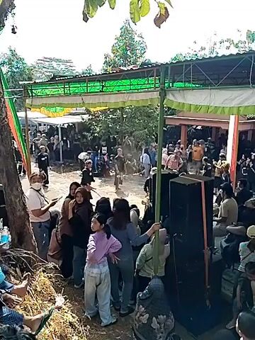 A crowd of people are gathered outdoors around a makeshift stage or performance area. Two performers are visible in the center of the frame, one wearing a black shirt with white lettering and a red and yellow mask with elaborate headwear, the other in a black outfit with a white patterned skirt. A third performer in a black shirt with white text is also visible. A blue barrel and what appears to be water are present near the stage. The ground is dirt and appears wet in some areas. Many spectators are standing, some are seated on the ground, and a few are visible in the foreground, looking towards the performance. A tent or canopy structure with green and white fabric is erected over the performance area. Trees and foliage are visible in the background, and sunlight suggests it is daytime. Speakers are positioned in the foreground, suggesting a musical or audio component to the event.