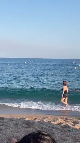 A woman in a black swimsuit walks into the ocean waves on a beach. The water is blue and there are some buoys in the distance. There is also another person swimming in the water to the right of the woman. The beach is made of dark sand and pebbles. The sky is clear and blue.