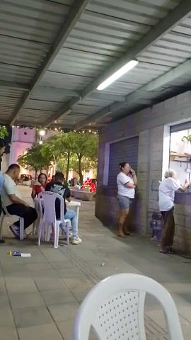 The camera moves through a narrow outdoor corridor. Groups of people sit in chairs under strings of lights. Some people are standing and talking to a person through a window. There are buildings on either side of the corridor.