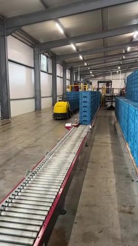 The video shows a brightly lit industrial warehouse with a concrete floor. On the left side of the frame, a long roller conveyor belt, with a red frame, extends into the scene. A small yellow automated guided vehicle (AGV) is positioned to the right of the conveyor belt. The AGV is transporting a stack of blue plastic crates, which are neatly arranged. The crates are moving along a pathway or track. To the right of the AGV and crates, a tall shelving unit, also filled with blue plastic crates, runs along the wall of the warehouse. The ceiling is high and features industrial lighting fixtures. The overall impression is of an automated warehouse system in operation. There are no people visible in the video.