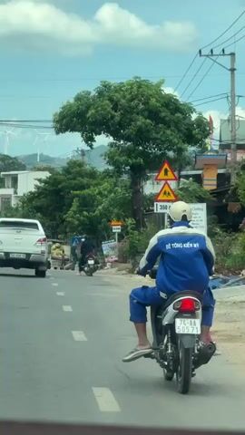 This media captures a scene in Lao Bảo Commune, Vietnam, likely during the day with a bright, cloudy sky. The primary subjects are vehicles on a paved road. A white pickup truck with a license plate reading "74C-030.18" is in the foreground, driving away from the camera. A motorcycle is also visible, with a person wearing a blue uniform and a white helmet riding it. The uniform has text on the back, which appears to be Vietnamese, possibly indicating a company name or profession.

The setting is a semi-rural or suburban area. Buildings, trees, and foliage line the sides of the road. In the background, a prominent feature is a cluster of wind turbines on a distant hill, suggesting the presence of renewable energy infrastructure. Traffic signs are visible, including one with a yellow triangle and a symbol that could indicate a hazard or a speed limit, and another sign that says "SLOW".

The activity appears to be ordinary road travel. The people on the vehicles are engaged in commuting or transportation. The overall mood is neutral, depicting a typical day in this location. The presence of the pickup truck, motorcycle, and shops suggests local commerce and daily life. The wind turbines are a significant visual element, highlighting a modern aspect of the landscape.
