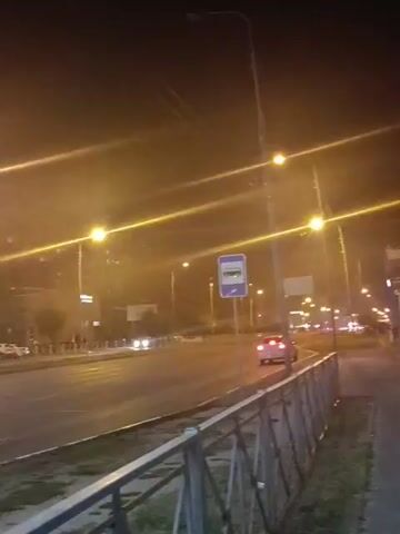 The video captures a nighttime scene on a city street. Streetlights illuminate the road and sidewalk, casting long glows. Several cars are visible on the road, some moving and some parked. A bus stop sign is present on the right side of the frame. Buildings line the street, with lights visible in some windows. The overall impression is of a quiet urban environment at night.