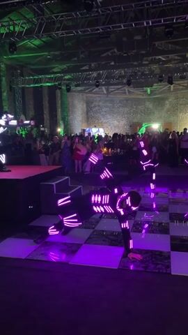A dance performance is taking place on a checkered floor illuminated with purple lights. Two performers in black suits adorned with neon pink lights are on the floor, one in a handstand pose and the other in a lower stance. In the background, a crowd of people is gathered, some seated at tables and others standing, watching the performance. The venue appears to be a large indoor space with exposed brick walls and industrial lighting.