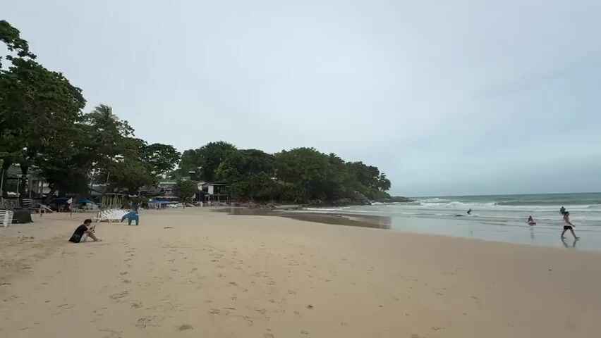 A wide shot of a beach on an overcast day. Sand dominates the foreground with footprints visible. In the middle ground, the shallow ocean waves gently lap against the shore. Several people are visible in the water, some wading, others swimming. Two individuals are walking along the wet sand at the water's edge. Further in the background, a small, tree-covered island is visible in the ocean. To the left of the frame, lush green trees line the edge of the beach, with a few small buildings and umbrellas nestled amongst them. A single person is seated on the sand to the far left, facing away from the camera. The sky is a uniform grey.