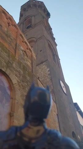 This is a low-angle shot of a towering, ancient church bell tower, made of stone and brick, with a clock face visible on its side. In the foreground, the back of a Batman figurine is prominently displayed, facing the bell tower. The figurine's dark cowl and ears are clearly visible, suggesting it's a toy or collectible.

The scene appears to be set in a European town, given the architectural style and the context of Peccioli, Italy. The clear blue sky in the background indicates it is daytime, likely late afternoon or early morning due to the warm light illuminating the brickwork of the church. There is no visible text in the media file. The juxtaposition of the stoic, weathered architecture with the iconic superhero figure creates an interesting, almost narrative, composition. The figurine's perspective suggests it is looking up at the imposing structure, perhaps contemplating its next move or simply observing the grandeur of the location.