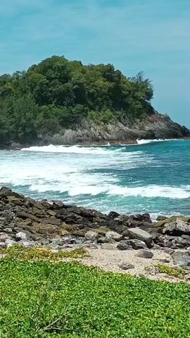 The video shows a rocky coastline with lush green vegetation in the foreground. In the middle ground, waves crash against a rocky shore. Further out, a small, tree-covered island juts out into the turquoise ocean, with more waves breaking around its base. The sky is a clear blue with a few wispy clouds. There are no people or animals visible in the video. The scene appears to be a natural landscape, likely a tropical or subtropical coastal environment.