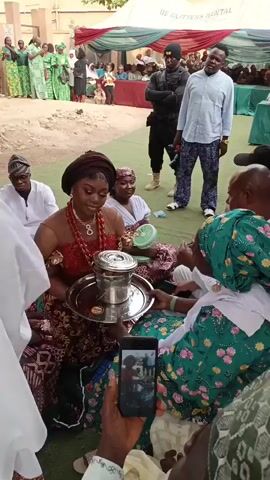 The video depicts an outdoor traditional ceremony or celebration, likely an engagement or wedding event, set in Sabon Gari West, Nigeria. The central figure is a young woman, possibly a bride, who is seated on the ground. She is dressed in vibrant traditional attire, featuring a red top with intricate patterns, a rich brown and gold patterned skirt, and a dark headwrap. She wears multiple strands of elaborate red beaded necklaces and bracelets, indicating significant cultural dress. Her face is bright with a joyful smile as she holds a silver tray with a lidded metal container, possibly a part of a ritual or presentation.

Another woman, dressed in a green floral-patterned dress and a matching headwrap, sits opposite her, engaged in interaction. Several other guests are also seated on the ground around them, observing the proceedings. A small portable fan is visible near one of the seated women, suggesting warm weather. In the immediate foreground, a person is holding a smartphone vertically, actively recording the event, while a microphone is partially visible in the upper left, indicating sound amplification or recording.

In the background, two men stand: one in a light blue shirt and patterned trousers, and another in a dark uniform, possibly security personnel. Behind them, under a large white tent, more guests are gathered, some standing and others seated. The tent has "DE GLITTERS RENTAL" printed on its side. Promotional posters are visible on a wall in the background, featuring images of women in traditional attire. The setting appears to be an open area with sandy ground, partially covered by green fabric, under bright daylight, contributing to the festive and celebratory atmosphere.