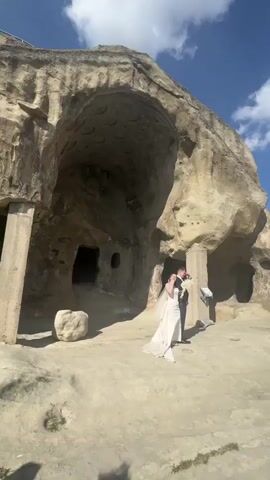 The video begins with a low-angle shot of a wedding photo shoot taking place near a large, natural rock formation with cave-like openings. A bride in a light-colored gown and groom in a dark suit pose while a photographer takes their picture. The camera then pans to the right, revealing a group of people standing along a railing, some taking photos with their phones. The view then transitions to a wide shot of a river valley, showcasing a flat, green landscape with a winding river and distant mountains under a sky with scattered clouds and a bright sun.