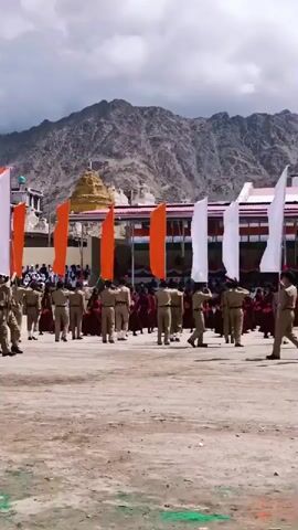 A group of people in uniform, likely cadets, are marching in formation outdoors. They are carrying flags of various colors including orange, white, and green. In the background, there are buildings and mountains. The ground appears to be dirt and gravel. The sky is partly cloudy.