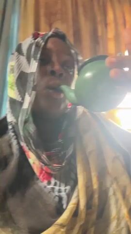 A woman wearing a black and white patterned headscarf with red flowers and a cream and yellow patterned scarf is drinking from a small green container with a spout. The container is being held up to her mouth by someone out of frame. The background includes beige curtains and a window. The lighting is bright, possibly from sunlight.