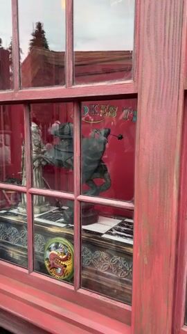 The video displays a vibrant shop window, likely situated in the Wizarding World of Harry Potter at Universal Studios Japan, given the Osaka, Japan context and thematic elements. The window features a rustic pinkish-red wooden frame with multiple glass panes.

Inside, against a red backdrop, an animated display is visible. Two knight statues on horseback are the central focus. The knight on the right is actively moving its arm up and down, simulating a jousting motion, accompanied by mechanical creaking sounds. The knight on the left remains static. To the far left, a static robed figure, possibly a queen or wizard, stands prominently. All figures are set upon ornate pedestals, some featuring black and white checkerboard patterns, evoking a chess game.

In the bottom center pane, a round, golden emblem with a red dragon design adds to the fantastical atmosphere. Reflections of people, trees, and surrounding architectural elements are visible on the window's surface, indicating it is daytime. Partially visible text inside the shop reads "JOKES T", suggesting a joke or novelty shop.