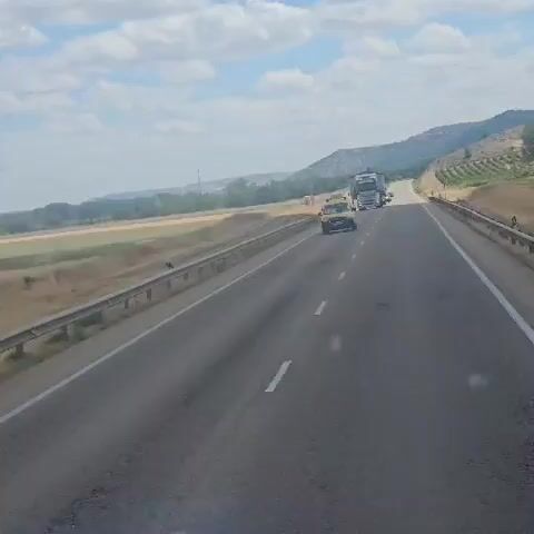 The video shows a view from inside a moving vehicle on a highway. A yellow van is seen in the foreground, followed by two large trucks. The first truck is white and appears to be transporting a large, box-like structure. The second truck is dark-colored and is carrying logs. The highway has multiple lanes and is bordered by a metal guardrail. To the left of the highway, there is a field with dry grass and a few trees in the distance. To the right, the landscape rises into a hilly terrain with some vegetation. The sky is partly cloudy. The video appears to be filmed during the daytime under sunny conditions.